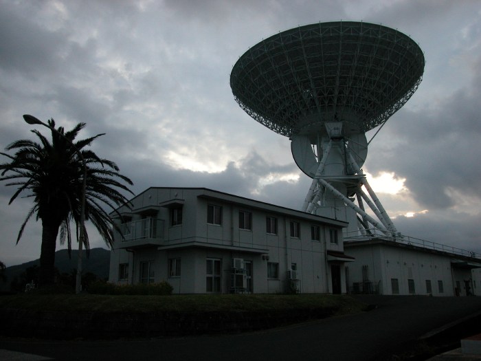 A palm tree anchors the end of a squat 2-story dormitory, while a large radio dish sits behind.  Sunlight pierces the low clouds to silhouette the scene. (63K JPEG)