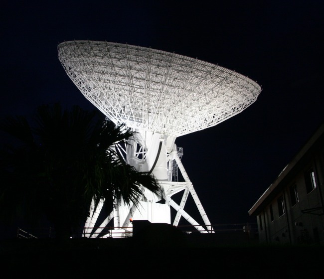 The spidery substructure of the parabolic dish sears into the blackness of night with the brightness of many million candlepower mercury lights, silhouetting the lazy fronds of a small palm foreground palm tree. (70K JPEG)