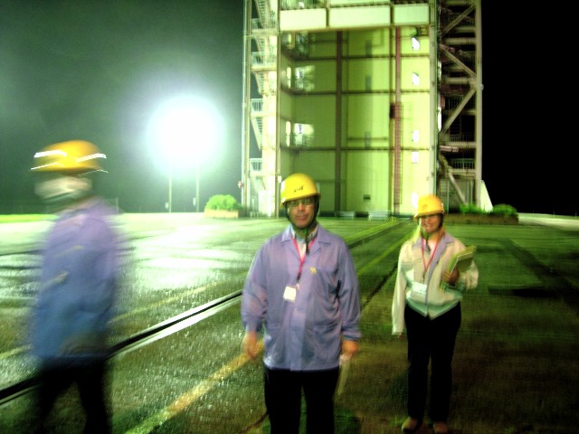 Distorted by motion blur in the dim greenish cast of mercury light, three people walk across the moist surface of the launch area, the tower standing patiently behind them. (73K JPEG)